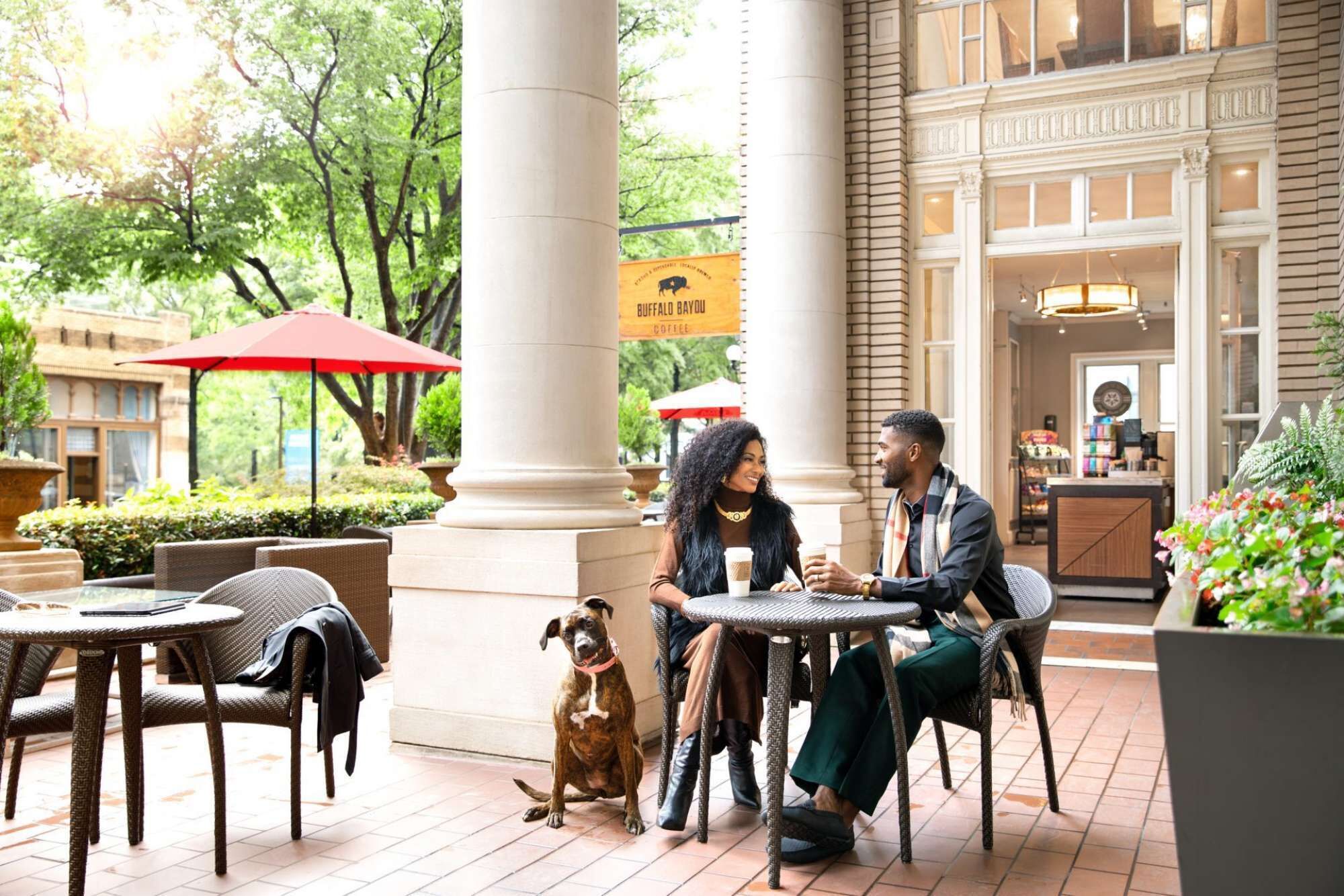 A couple dining outdoors at a cafe with a dog, surrounded by wicker furniture and a large column.