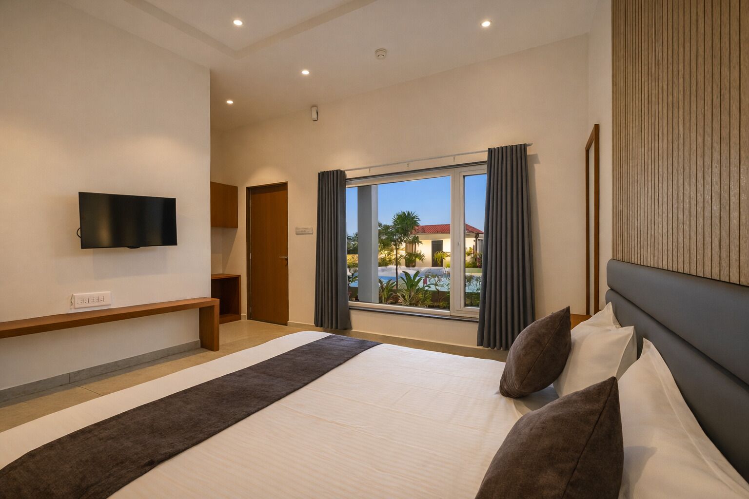 A hotel room with a bed, a TV mounted on the wall, a window with curtains, and a view of a pool and buildings outside.