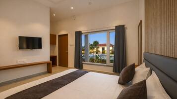 A hotel room with a bed, a TV mounted on the wall, a window with curtains, and a view of a pool and buildings outside.
