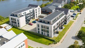 Ein modernes Wohngebäude mit mehreren Etagen, einem Parkplatz und einer angrenzenden Wasserfläche.