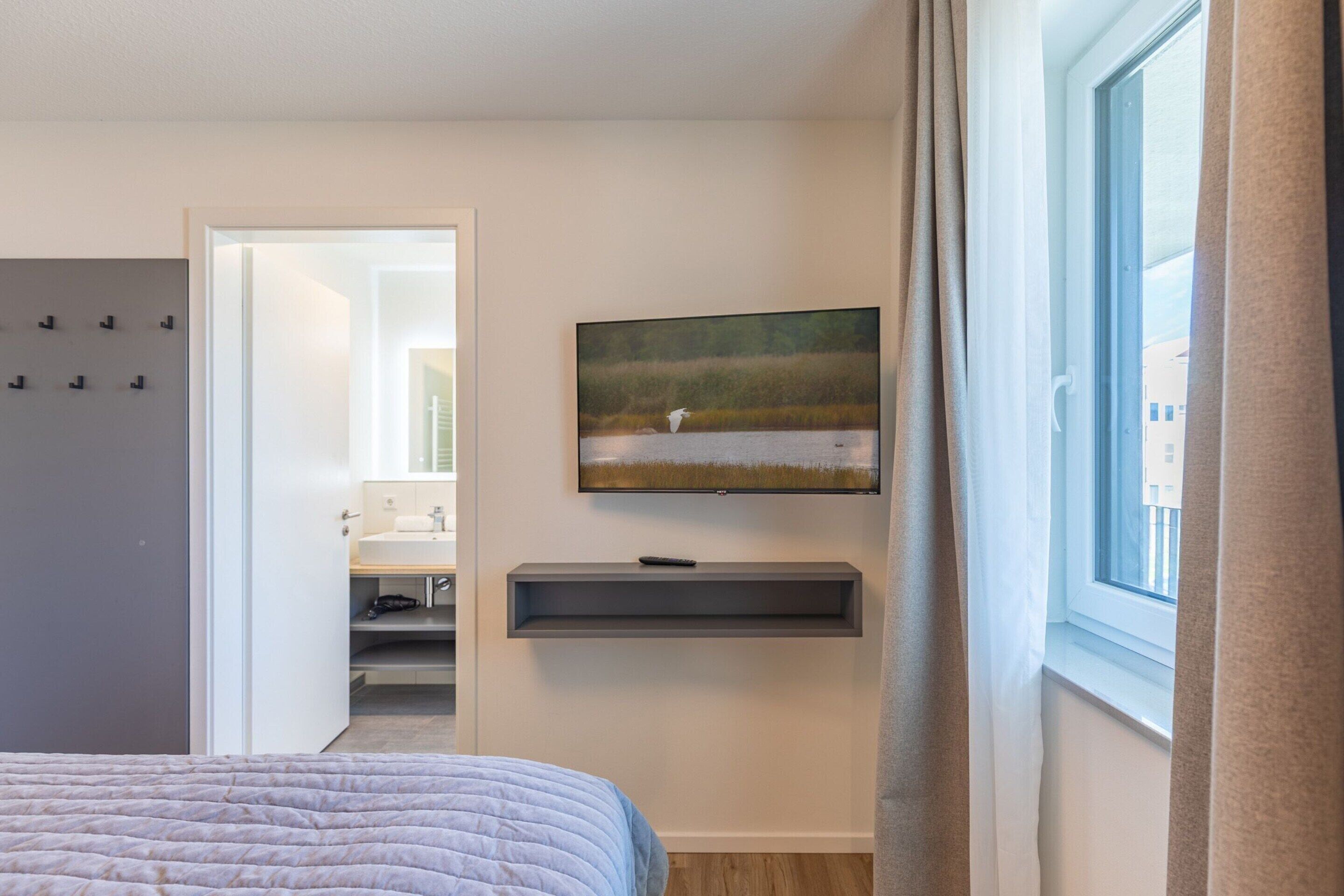 A bedroom with a bed, a TV mounted on the wall, a window with curtains, and a bathroom visible through an open door.