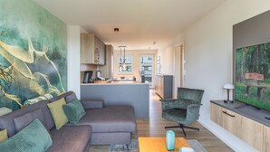 Ein modernes Wohnzimmer mit einer Couch, einem Couchtisch und einem Fernseher. Im Hintergrund ist eine Küche mit Essbereich zu sehen.