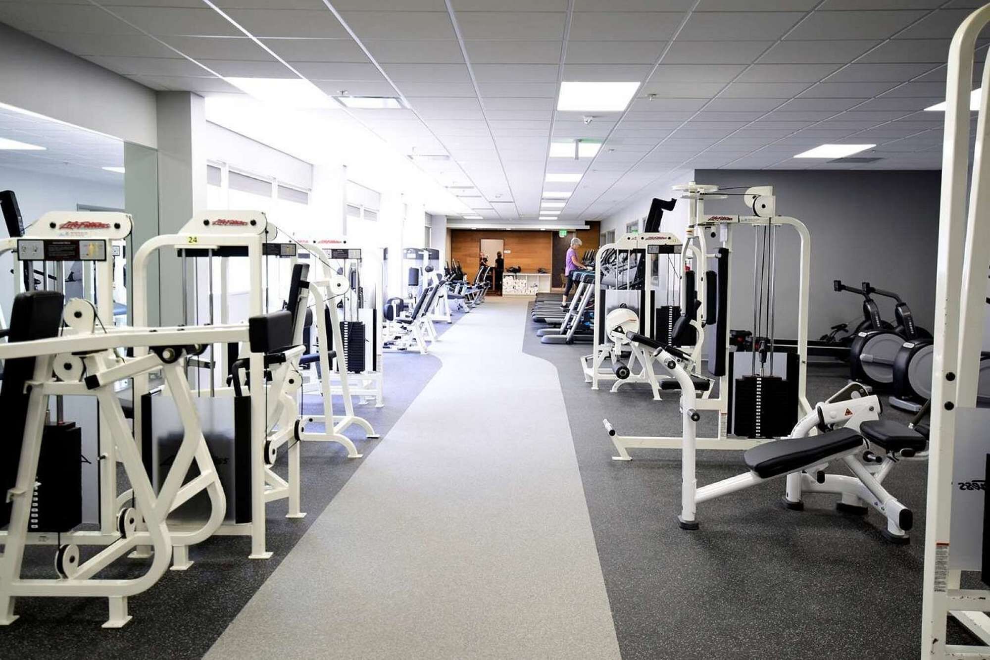A gym with various exercise machines, a central walkway, and a reception area in the background.