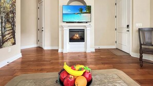 Un salon avec une cheminée, un téléviseur fixé au-dessus et un panier de fruits posé sur le sol.