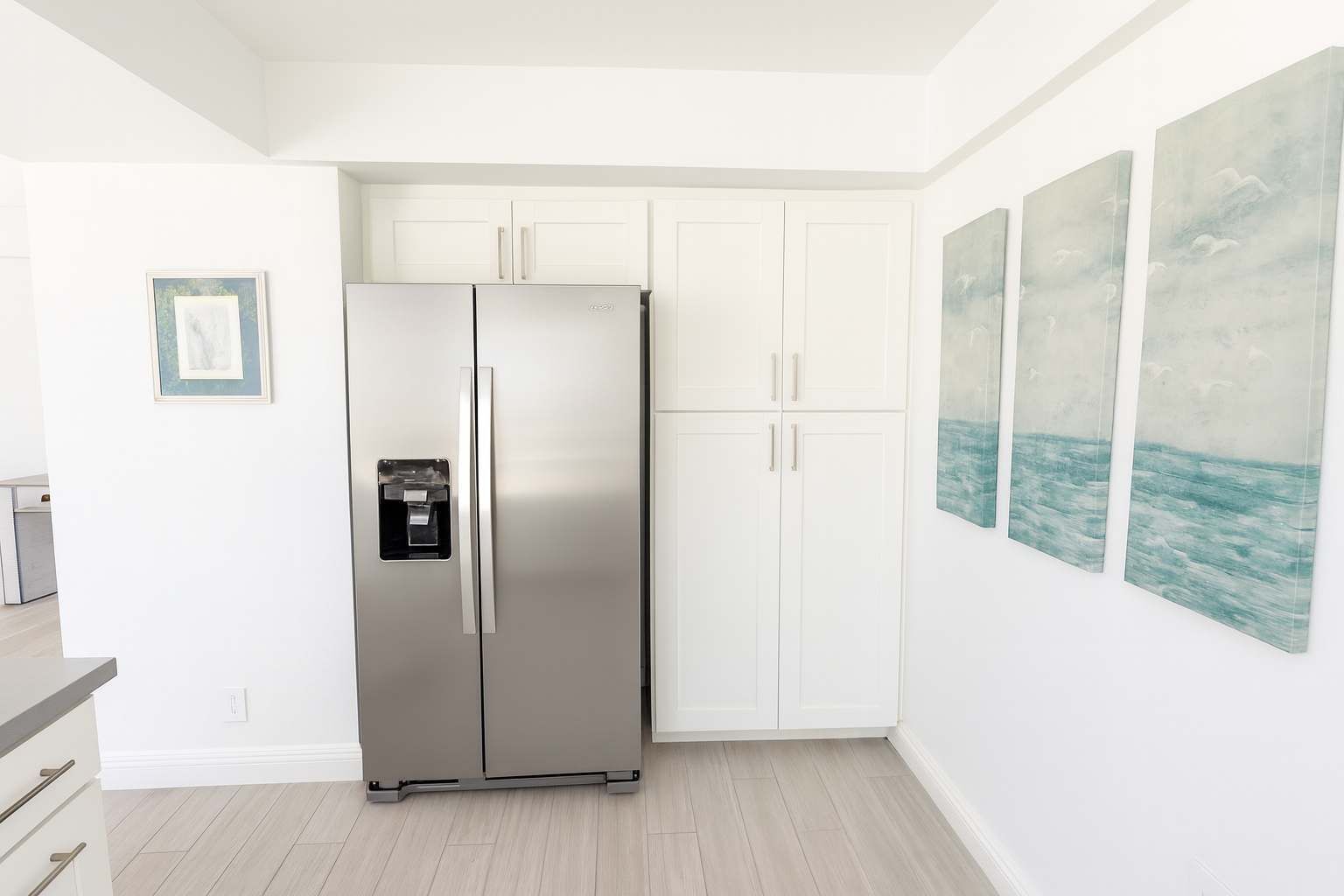 A modern kitchen with a stainless steel refrigerator, white cabinetry, and a framed picture on the wall.