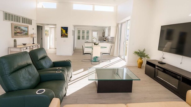 Ein modernes Wohnzimmer mit einem Glastisch, einer Couch, einem Fernseher und einer Küche im Hintergrund.
