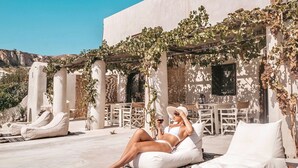 Een zonnig terras buiten met witte loungestoelen en een pergola begroeid met wijnranken.