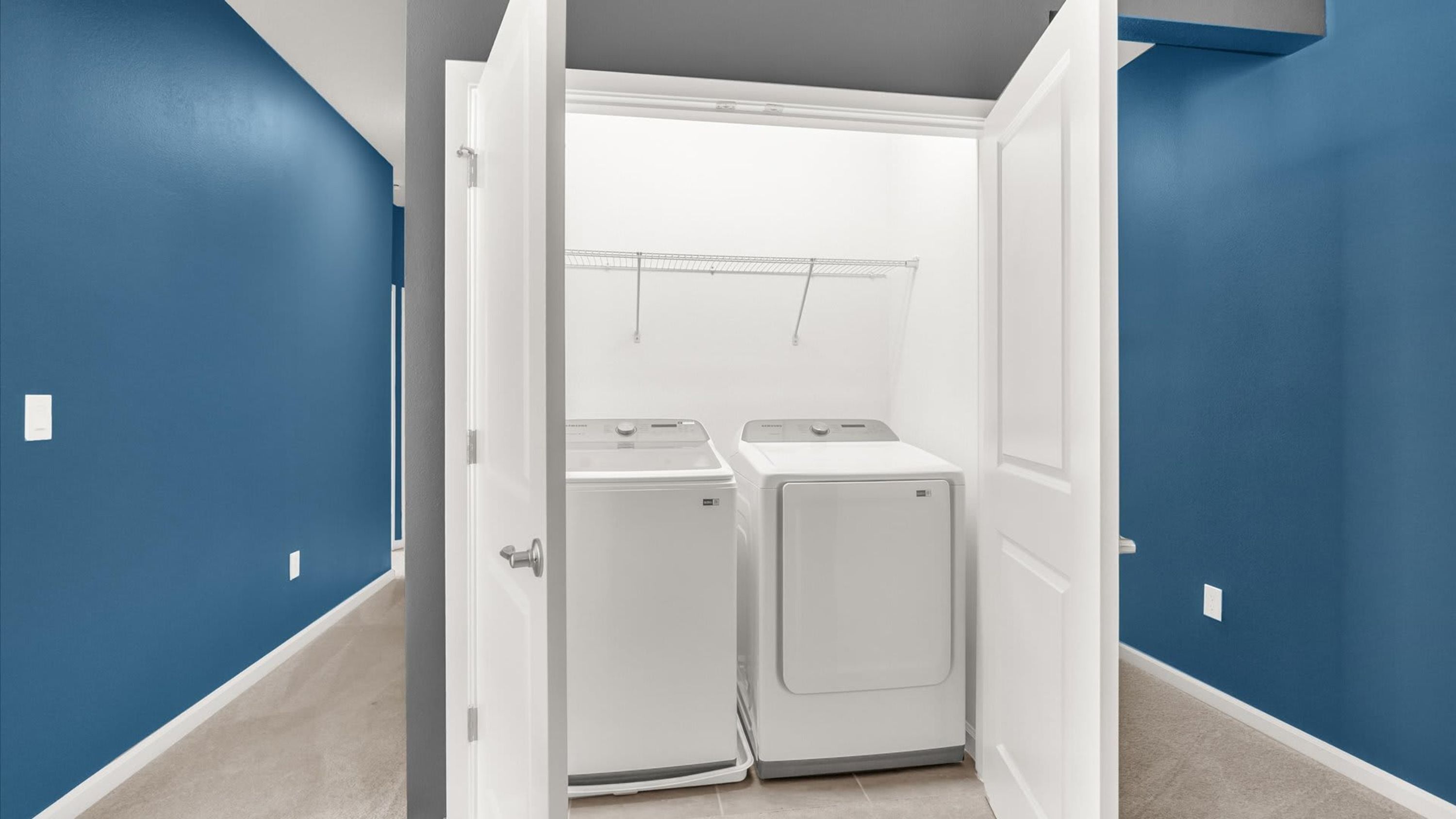 A laundry room with two white washers and dryers, a metal rack above the dryers, and a door leading to another room.