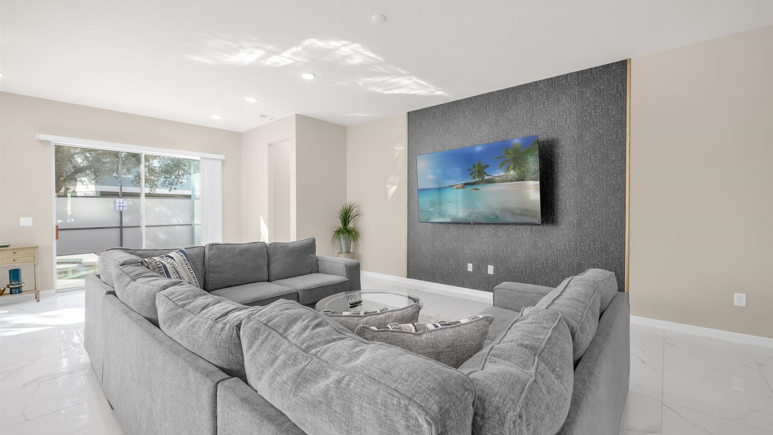 A modern living room with a large gray sectional sofa, a coffee table, and a flat-screen TV mounted on a textured gray wall.
