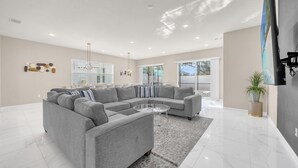 A modern living room with a grey sectional sofa, a glass coffee table, and a large window providing natural light. - Upstay Spacious Villa w Pool Hot Tub Game Room (Kissimmee)
