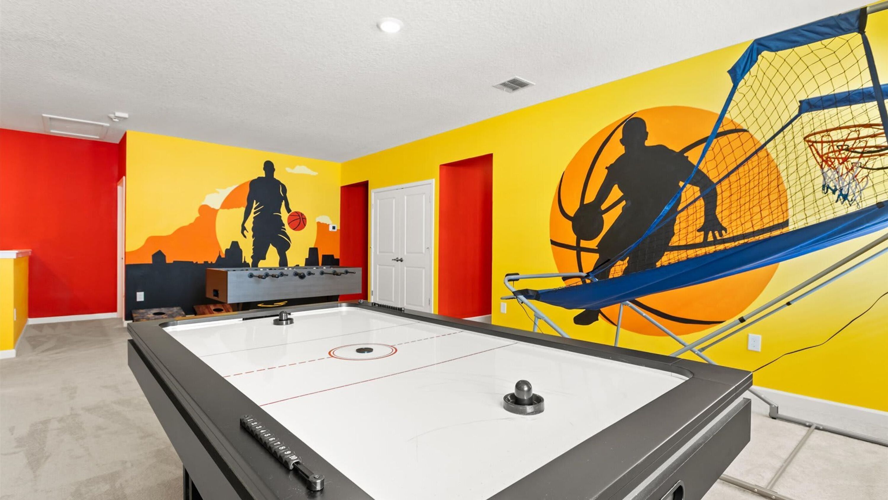 Una habitación con mesa de fútbol de mesa, aro de baloncesto y un mural con motivos de baloncesto.