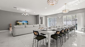 A modern dining room with a long table set for a meal, a sofa, and a large window providing natural light. - Upstay Veranda Palms Villa Pool Hot Tub Games (Kissimmee)