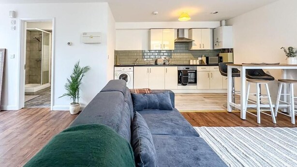 A modern apartment with a living area featuring a grey sofa, a small kitchen with white cabinets, and a dining area with stools. - 58A Thorpe Road, Norwich (Norwich)