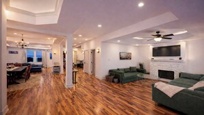 Uma sala de estar ampla, com assoalho de madeira, um sofá verde, lareira e ventilador de teto.