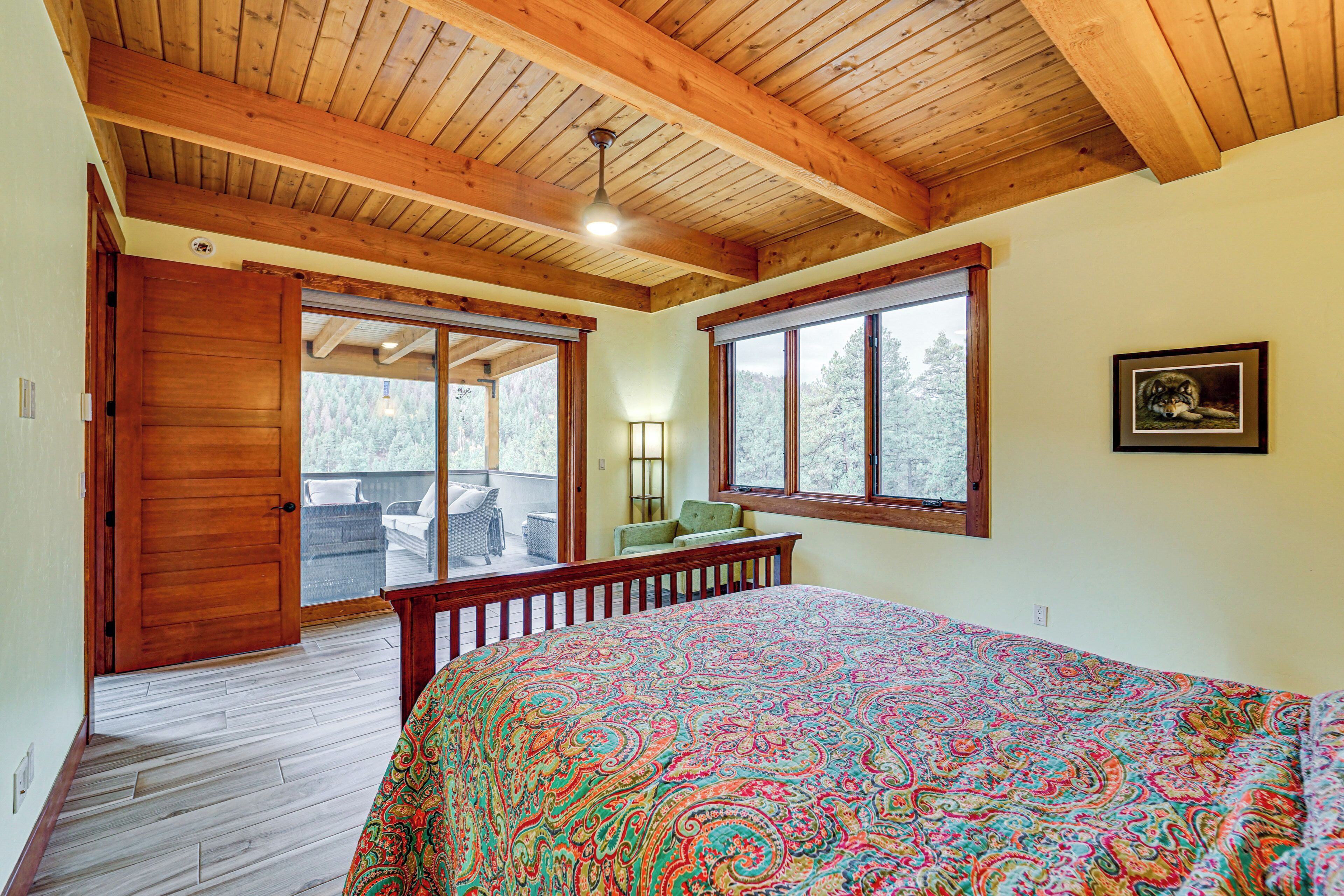Une chambre avec un plafond en bois, un grand lit recouvert d’une courtepointe colorée, une fenêtre donnant sur les arbres et une porte menant à un balcon.