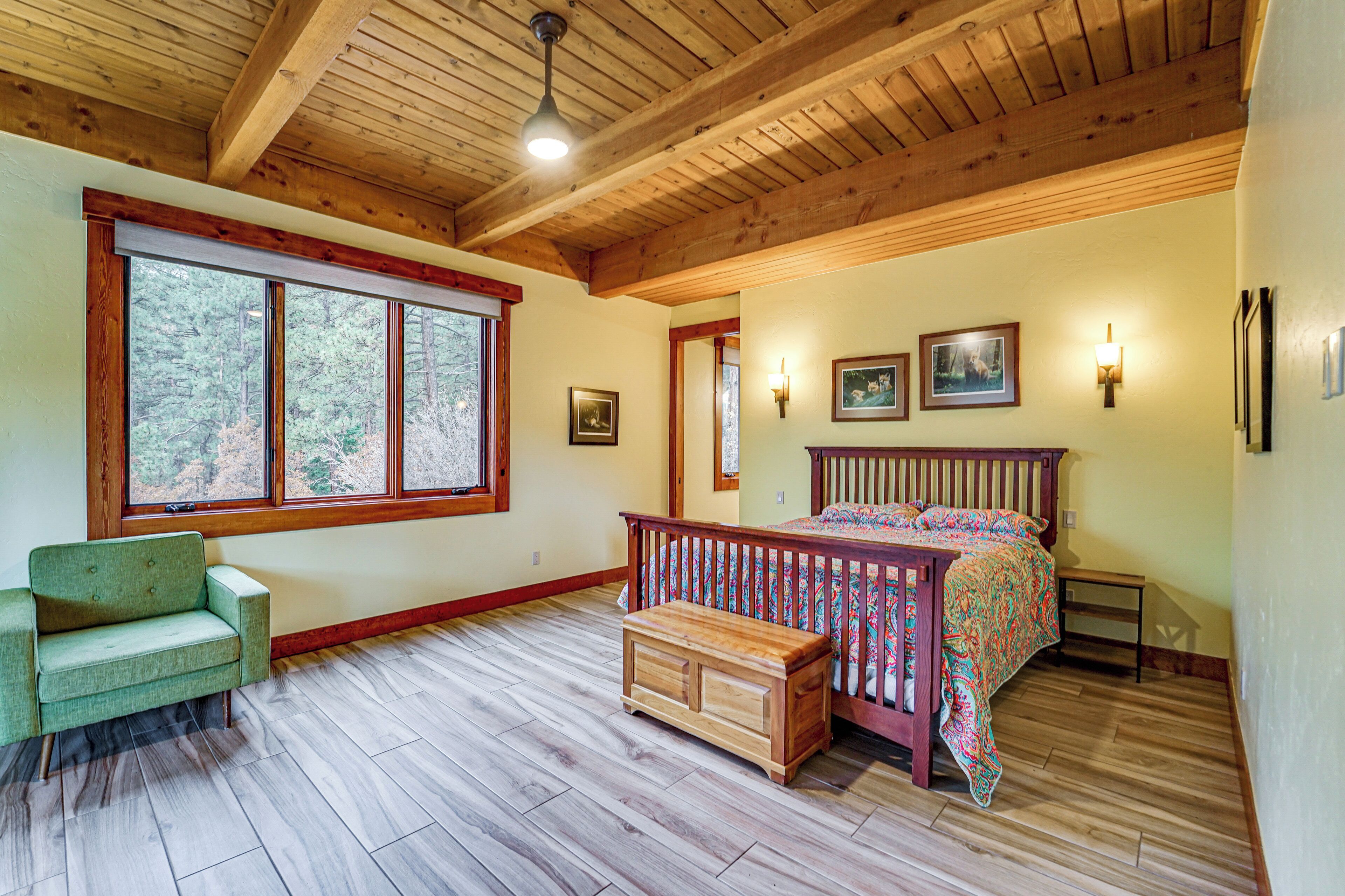 A bedroom with a wooden ceiling, a large window, a bed, a bench, and wall-mounted lights.