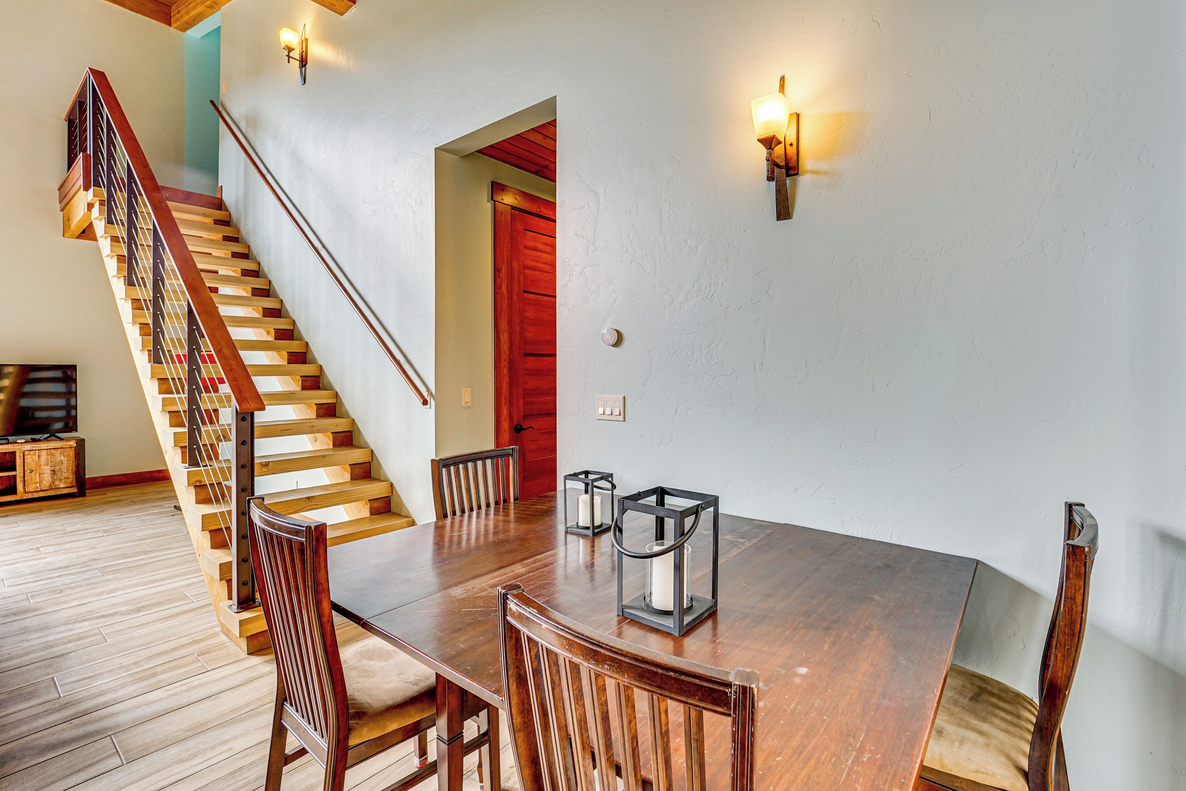 Una escalera de madera con barandilla de metal, una mesa de comedor con sillas y lámparas fijadas a la pared.