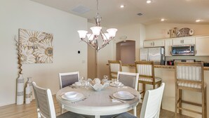 A dining area with a round table set for four, surrounded by white chairs with grey cushions. A chandelier hangs above the table, and a decorative wall piece is visible to the left. - Quiet Escape in The Villages! House w/ Lanai (Wildwood)