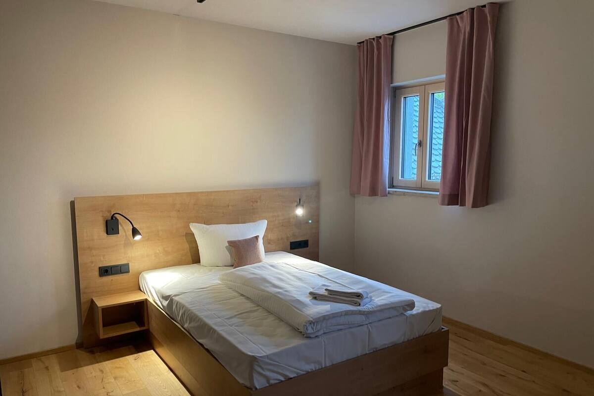 Et soveværelse med sengegavl i træ, et natbord med lampe, et vindue med gardiner og en pænt redt seng.