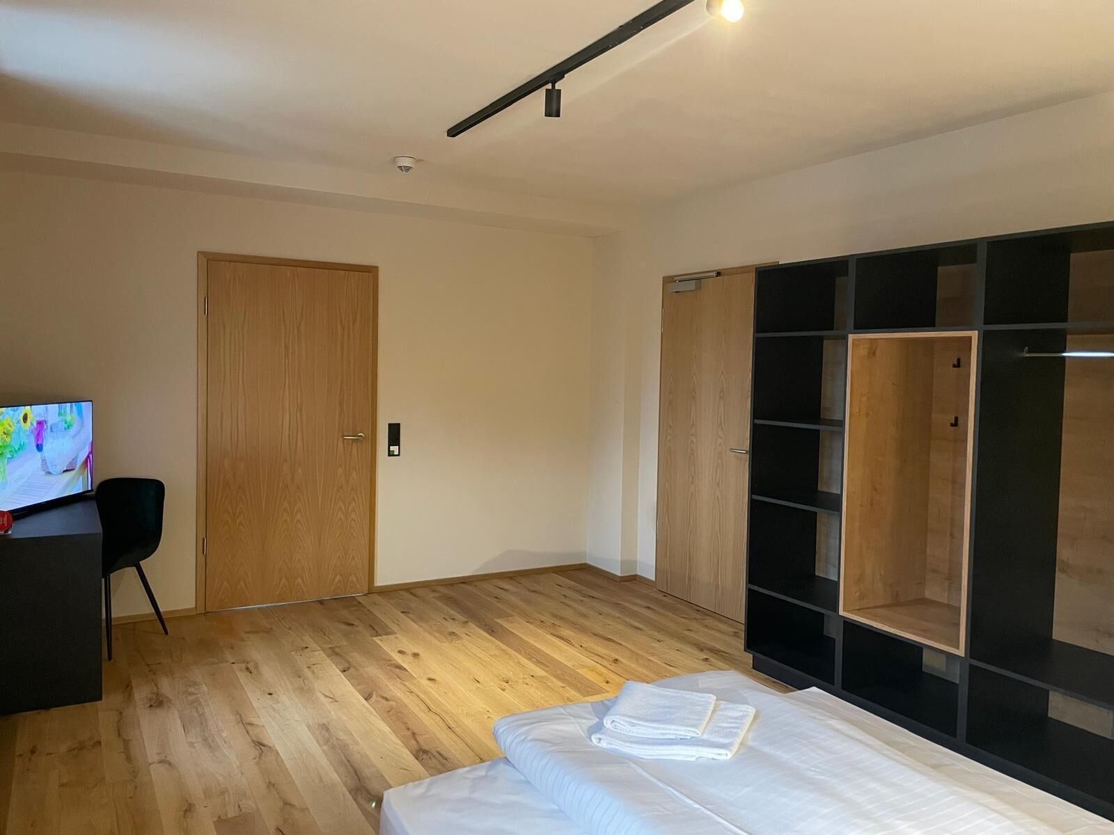Un dormitorio moderno con suelo de madera, una cama con ropa blanca, un televisor sobre un escritorio y un armario con paneles negros y de madera.