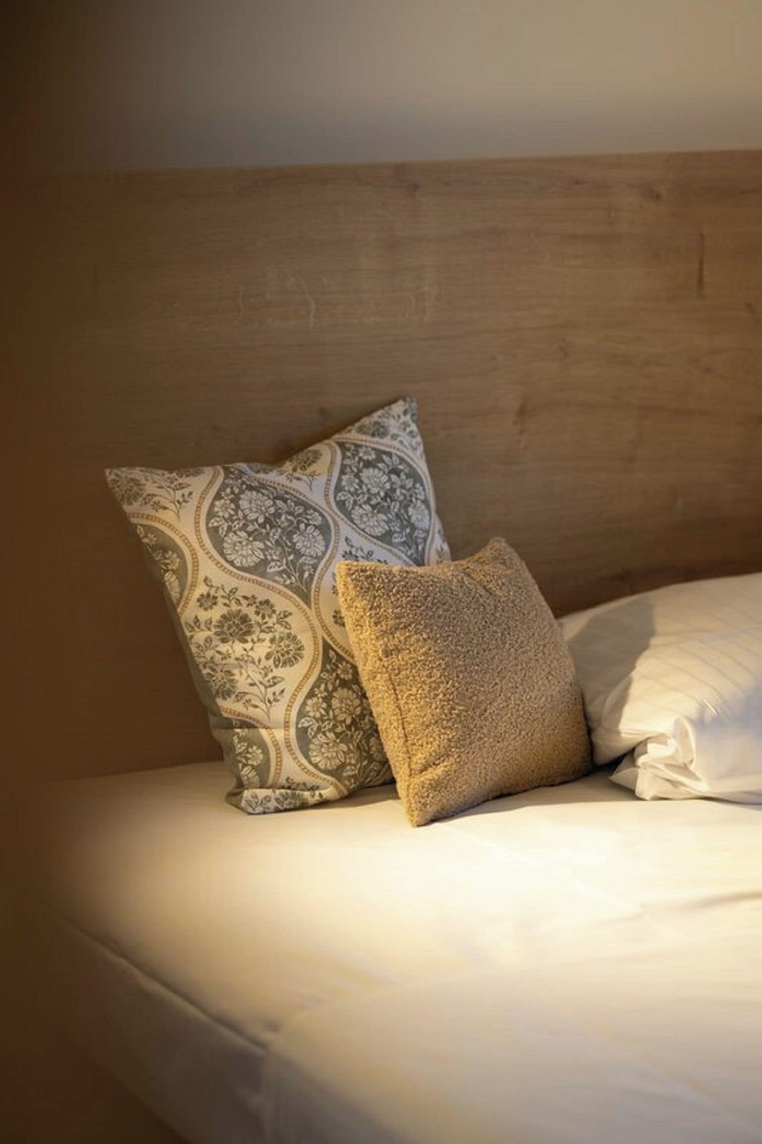 Een bed met een houten hoofdbord, twee kussens met een patroon, en een effen wit kussen.