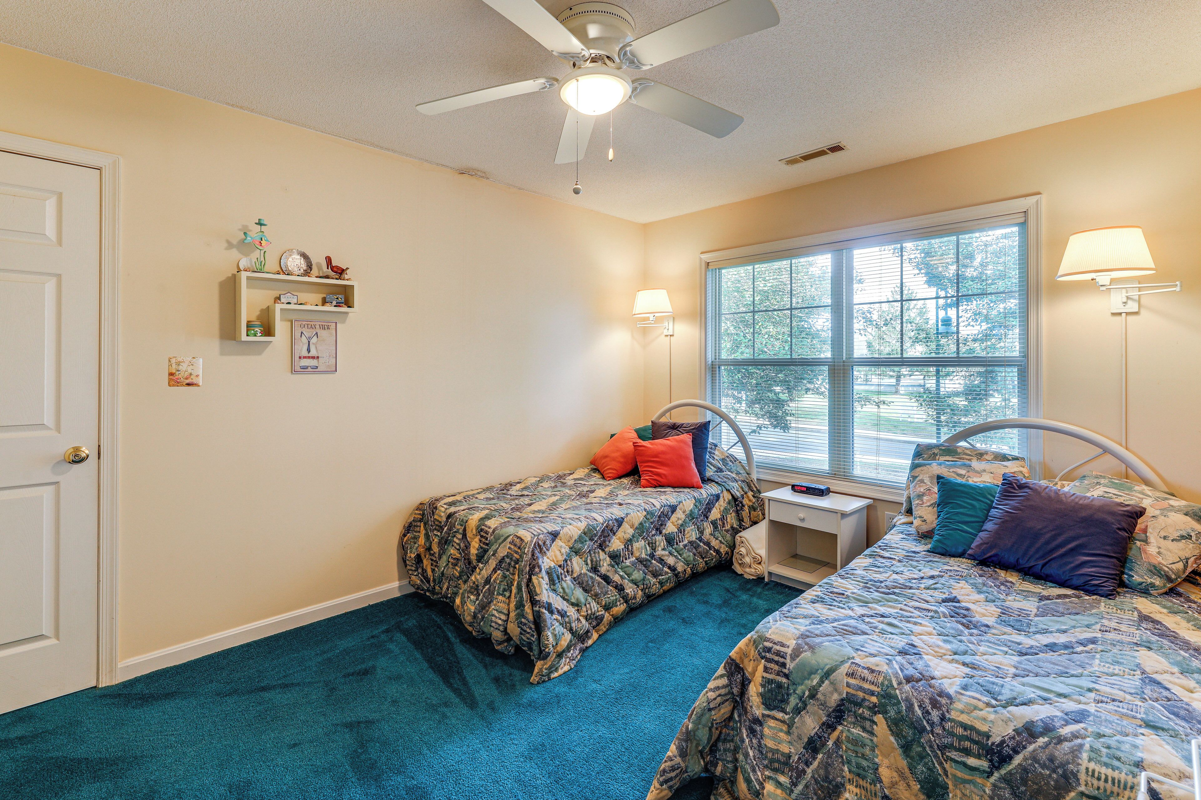 A bedroom with two beds, a ceiling fan, a window with blinds, and a nightstand with a lamp.