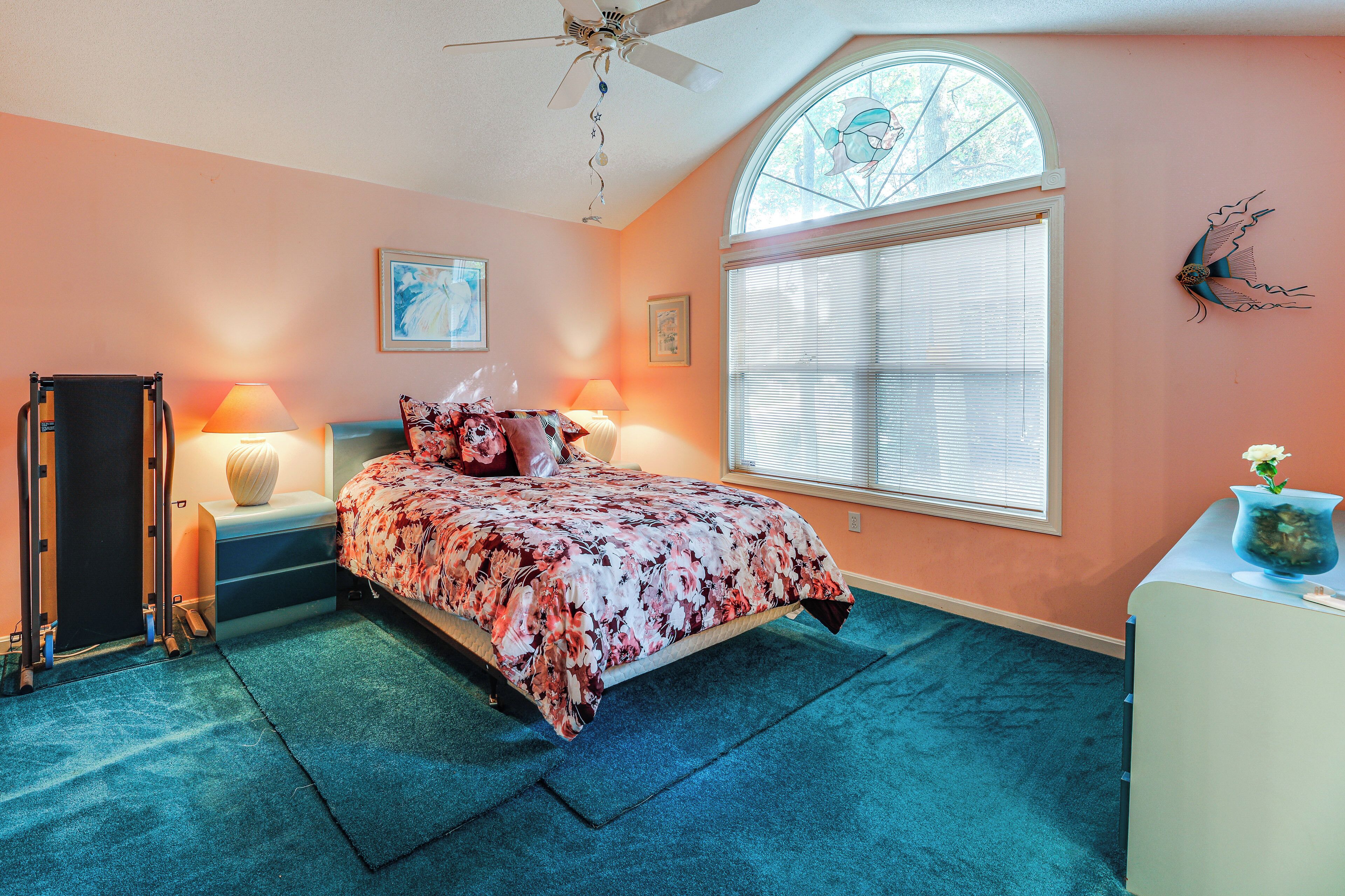 A bedroom with a bed, a desk, a chair, a ceiling fan, a window, and a wall decoration.