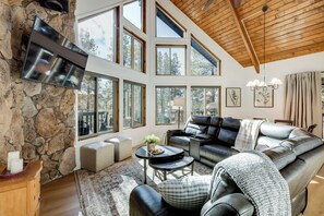 A spacious living room with a stone wall, large windows, a leather sofa, and a flat-screen TV mounted on the wall. - Family Getaway w/ Hot Tub! 20 Mi to Flagstaff (Munds Park)