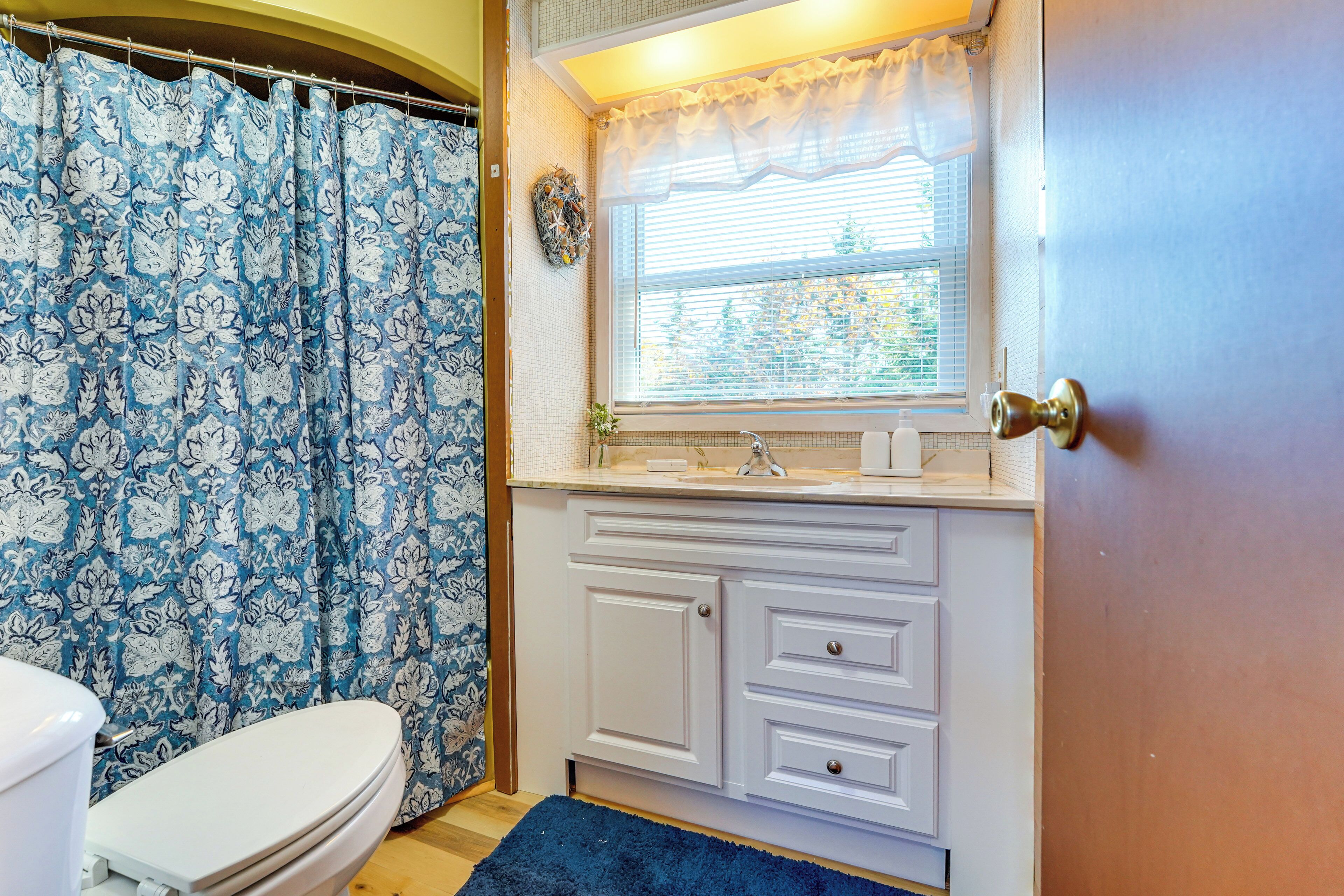 Une salle de bain avec des toilettes, un lavabo placé sous une fenêtre et un rideau de douche orné d’un motif floral.