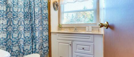 Une salle de bain avec des toilettes, un lavabo placé sous une fenêtre et un rideau de douche orné d’un motif floral.
