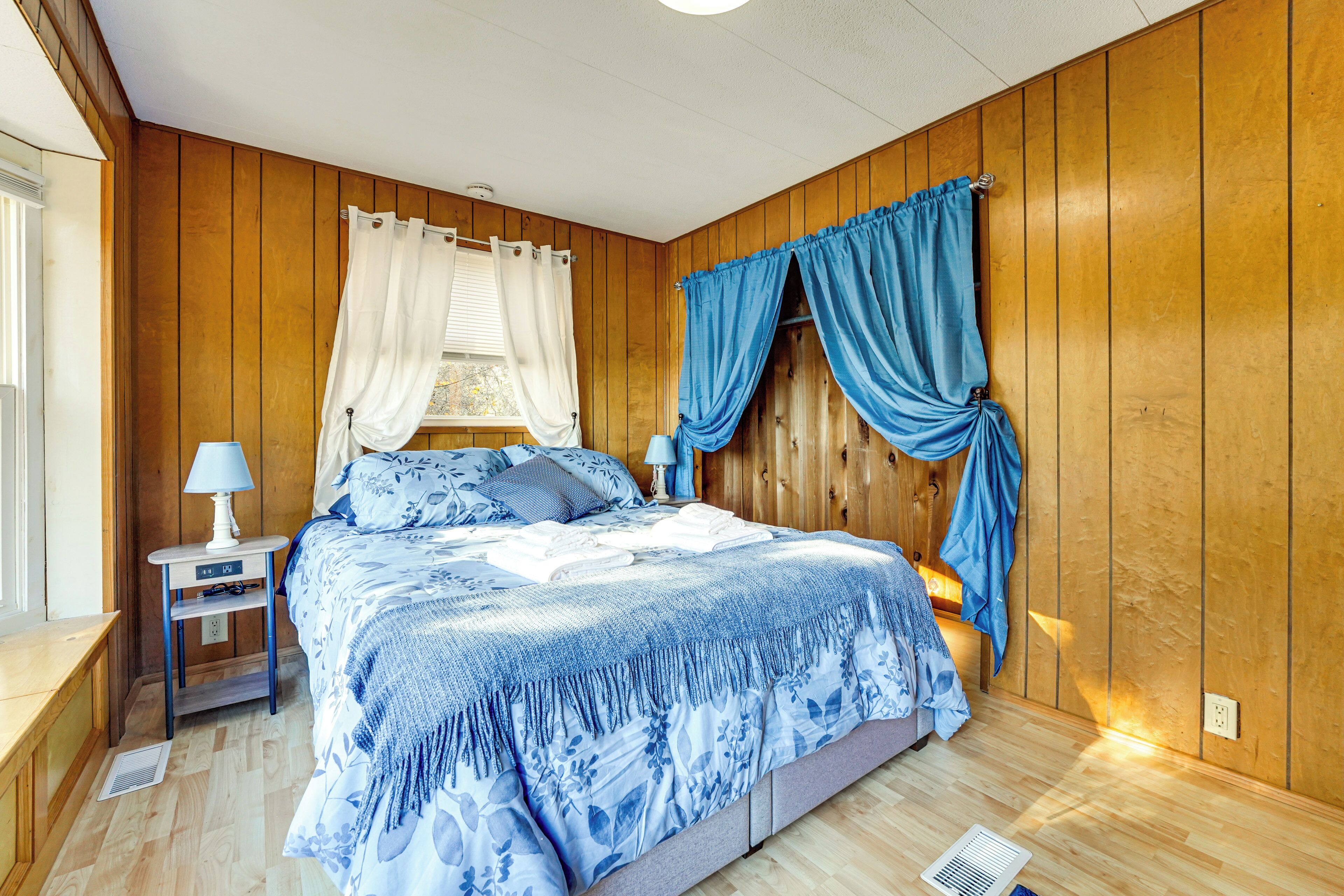 Una camera da letto con pannelli in legno, un letto con copripiumino a fiori blu, due comodini con lampade e una finestra con tende bianche.