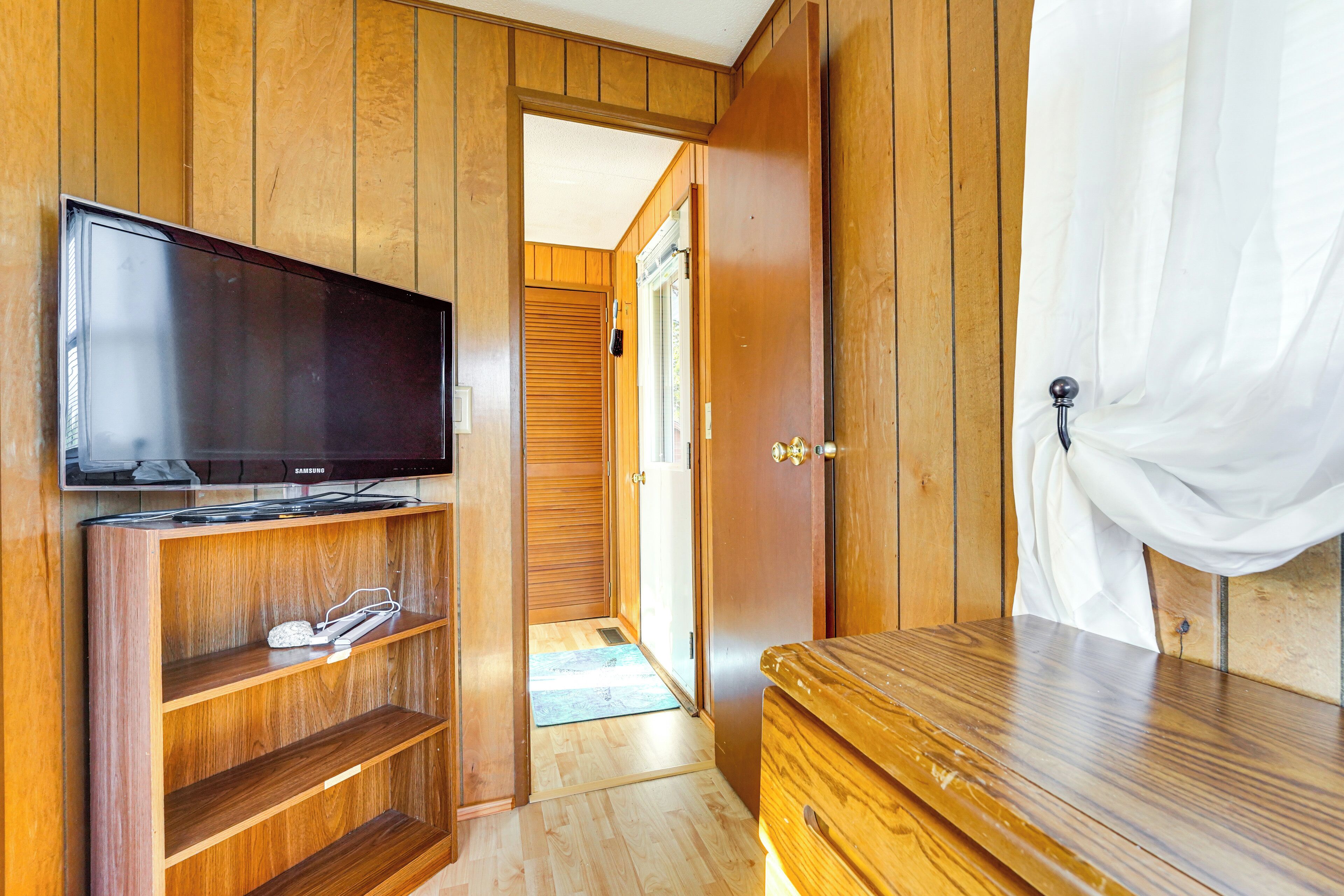 A wooden-paneled room with a television on a shelf, a door leading to another room, and a white curtain on a window.