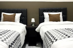 A hotel room with two beds, each with a black headboard and white bedding. There is a nightstand between the beds with a lamp on it. - Griya Inn (Tembalang)
