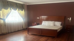 A bedroom with a large bed, wooden flooring, a nightstand, and a window with curtains.
