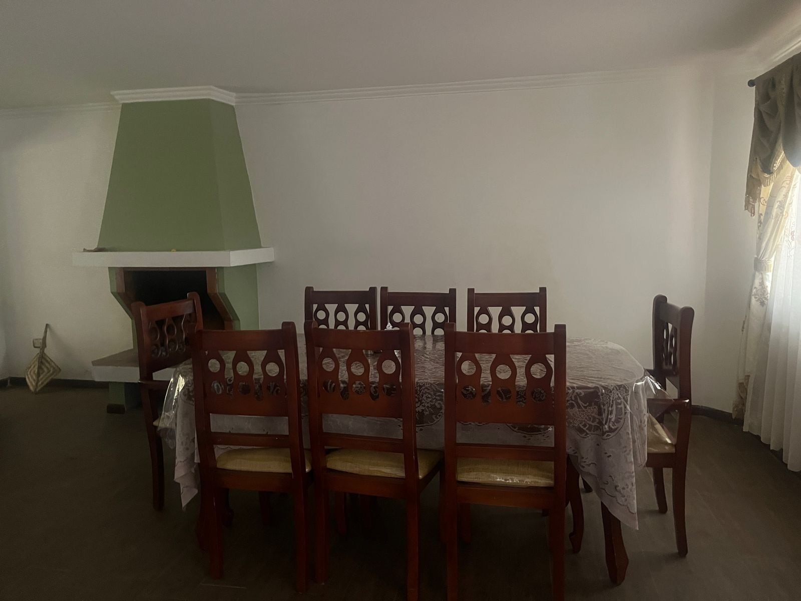 A dining area with a table set for six, surrounded by wooden chairs with intricate back designs. A green lamp is positioned to the left of the table, and a white tablecloth covers the table.