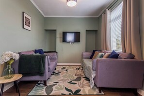 Een woonkamer met twee paarse banken, een flatscreen televisie, een kamerbreed tapijt met een patroon en een vaas met bloemen.