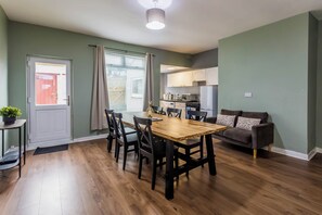 Een moderne keuken met witte kastjes, een eethoek met een houten tafel en stoelen, en een grijze bank.