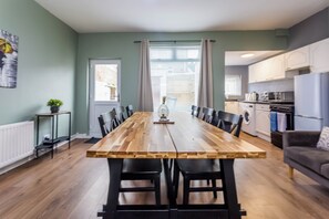 A dining area with a wooden table and chairs, a kitchen in the background, and a view through a window with curtains. - Toast Stays - The Northern View (Sherburn Hill)