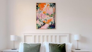 Un lit bien fait, avec une literie blanche et deux coussins verts, flanqué de deux tables de chevet blanches surmontées de lampes, dans une chambre dotée d’une tête de lit blanche et d’un tableau abstrait coloré au-dessus du lit.