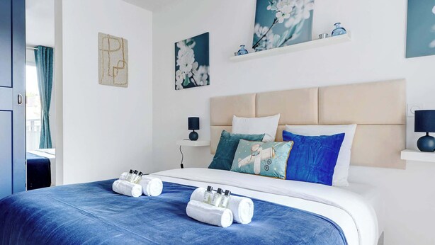 Hotellihuone, jossa on suuri sänky, siniset vuodevaatteet ja pääty. Sängyn molemmilla puolilla on yöpöydät, joissa on lamput, ja sängyn yläpuolella on pieni hylly.