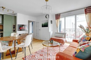 A modern living room with a wooden dining table, white chairs, a glass coffee table, and a sofa with colorful cushions. A chandelier hangs from the ceiling, and there is a large window with curtains. - Appartement avec balcon – Proche de l’Hippodrome (Suresnes)