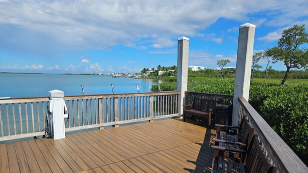 Eine Holzterrasse mit Geländer und Blick auf ein Gewässer, eine Bank und ein Grill.