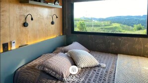 A bedroom with a bed, pillows, a shelf with decorative items, wall-mounted lamps, and a window with a view of green hills. - Via Kairos - Rancho Queimado - SC (Rancho Queimado)