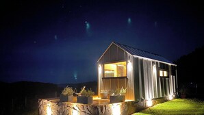 A modern, illuminated house with a stone foundation and a wooden deck, set against a night sky with visible stars. - Via Kairos - Rancho Queimado - SC (Rancho Queimado)