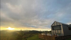 A rustic building with a stone foundation and metal siding, situated in a grassy field with a sunset in the background. - Via Kairos - Rancho Queimado - SC (Rancho Queimado)