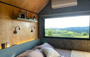 A bedroom with a bed, pillows, a shelf with decorative items, wall-mounted lamps, and a window with a view of green hills. - Via Kairos - Rancho Queimado - SC (Rancho Queimado)