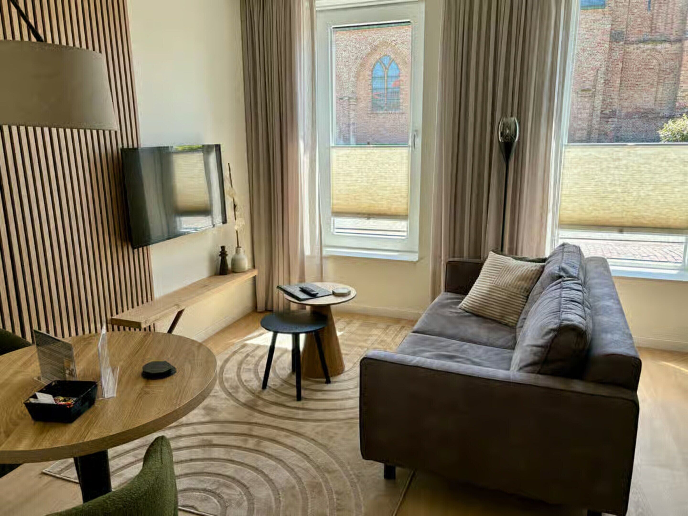 Een moderne woonkamer met een grijze bank, een ronde houten salontafel, een aan de muur bevestigde televisie en een groot raam met uitzicht op een bakstenen gebouw.
