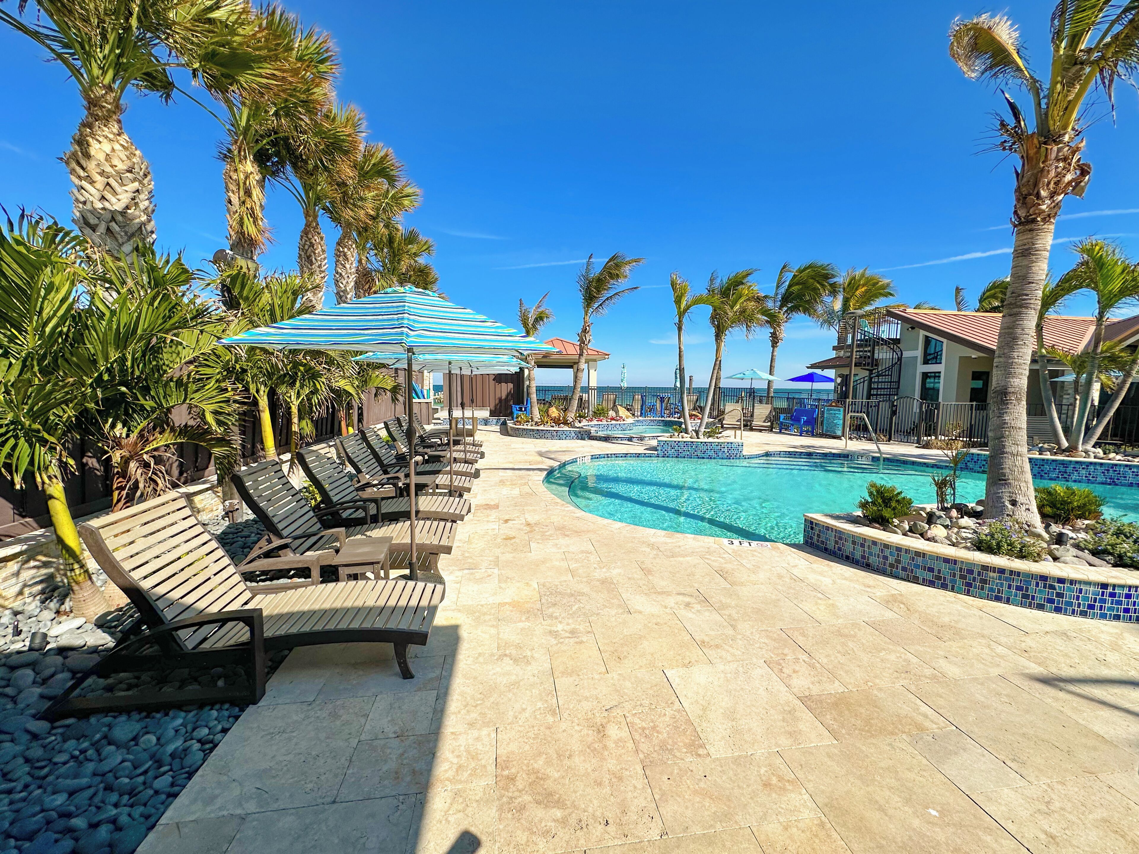 A poolside area with lounge chairs and umbrellas, a clear blue sky, and a view of the ocean.