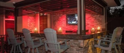 Un espace bar extérieur avec des fauteuils Adirondack, un mur en pierre et un téléviseur fixé au mur.