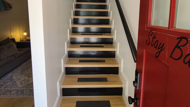 A staircase with alternating black and wooden steps, a red door with a 'Stay Back' sign, and a framed picture collection on the wall. - Barefoot Cottage A Quiet Coastal Retreat (annapolis)